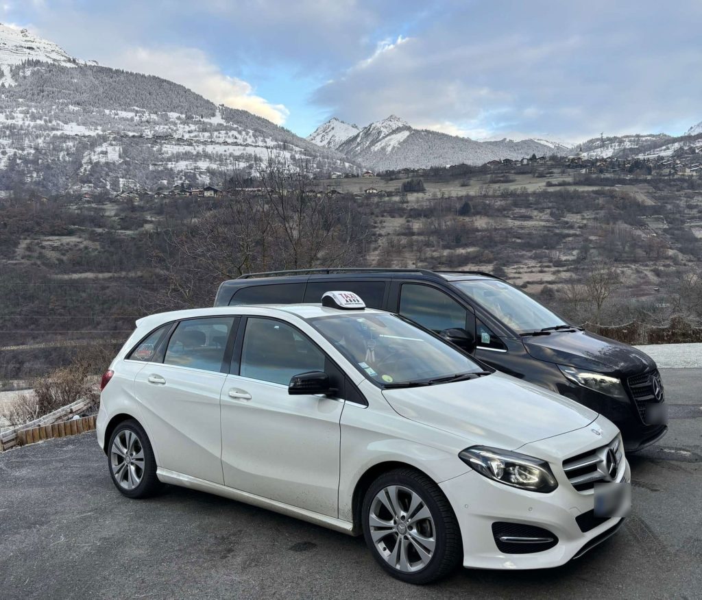 Photo de deux véhicules de l'entreprise taxi Mademoiselle prise sur le coté, une voiture blanche à l'avant, avec l'insigne de taxi sur le toit, et une autre voiture à l'arriere, un van noir. Les deux véhicules sont garés sur un parking avec en arrière plan une montagne avec un peu de neige, et un ciel plein de nuage et soleil taxi