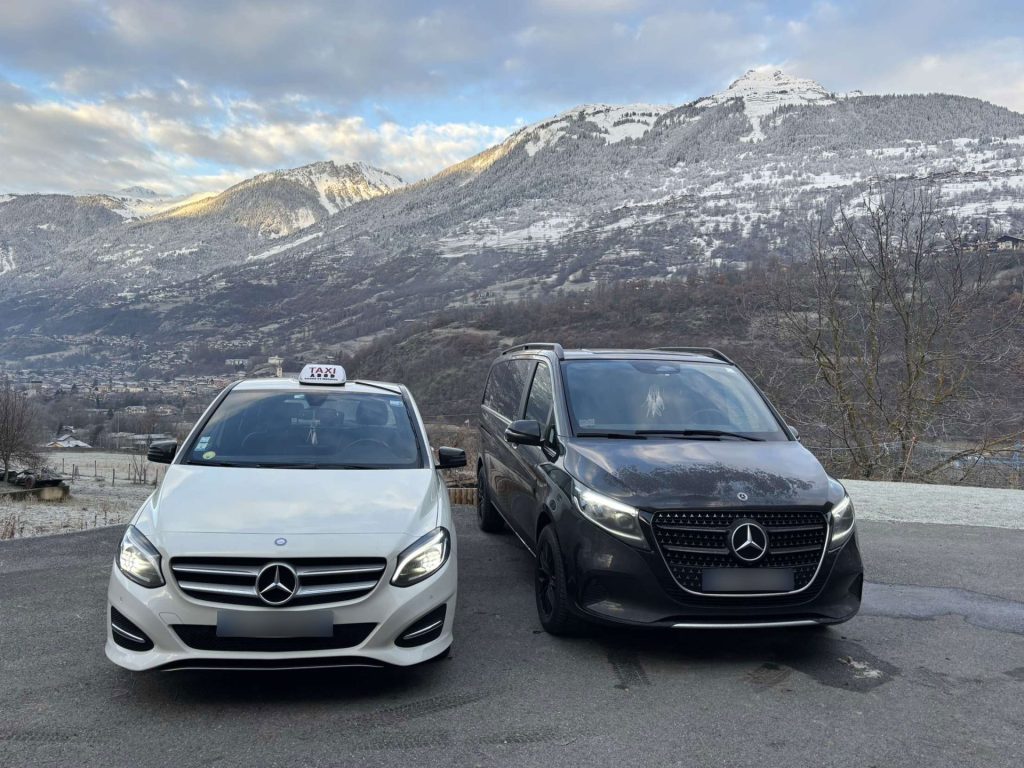 Photo de deux véhicules de l'entreprise taxi Mademoiselle prise de devant, une voiture blanche à gauche, avec l'insigne de taxi sur le toit, et une autre voiture à droite, un van noir. Les deux véhicules sont garés sur un parking avec en arrière plan une montagne avec un peu de neige, et un ciel plein de nuage et soleil taxi